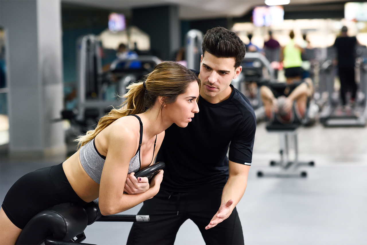 Personal trainer helping young woman lift weights while working out in a gym